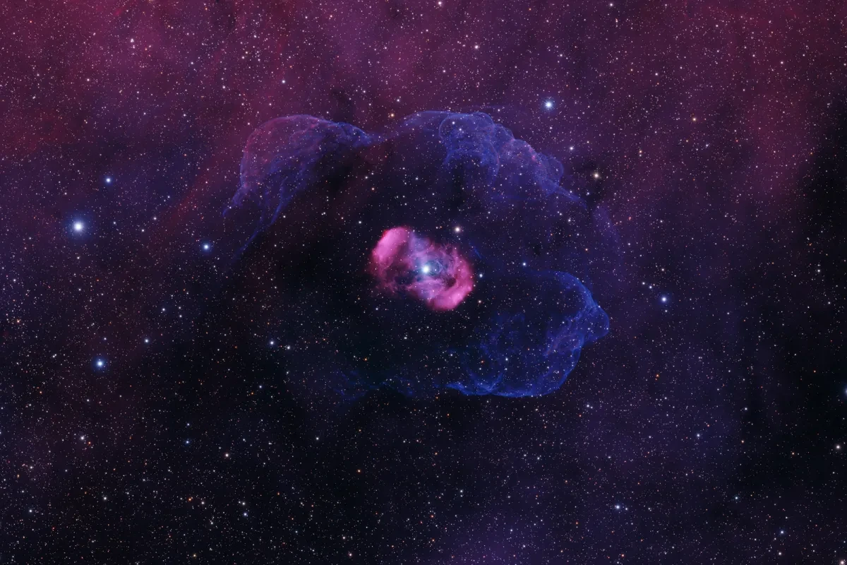 Bright star in the centre of a small egg-shaped pink nebula surrounded by protrusions of purple nebulae clouds, against a deep purple sky smattered with stars