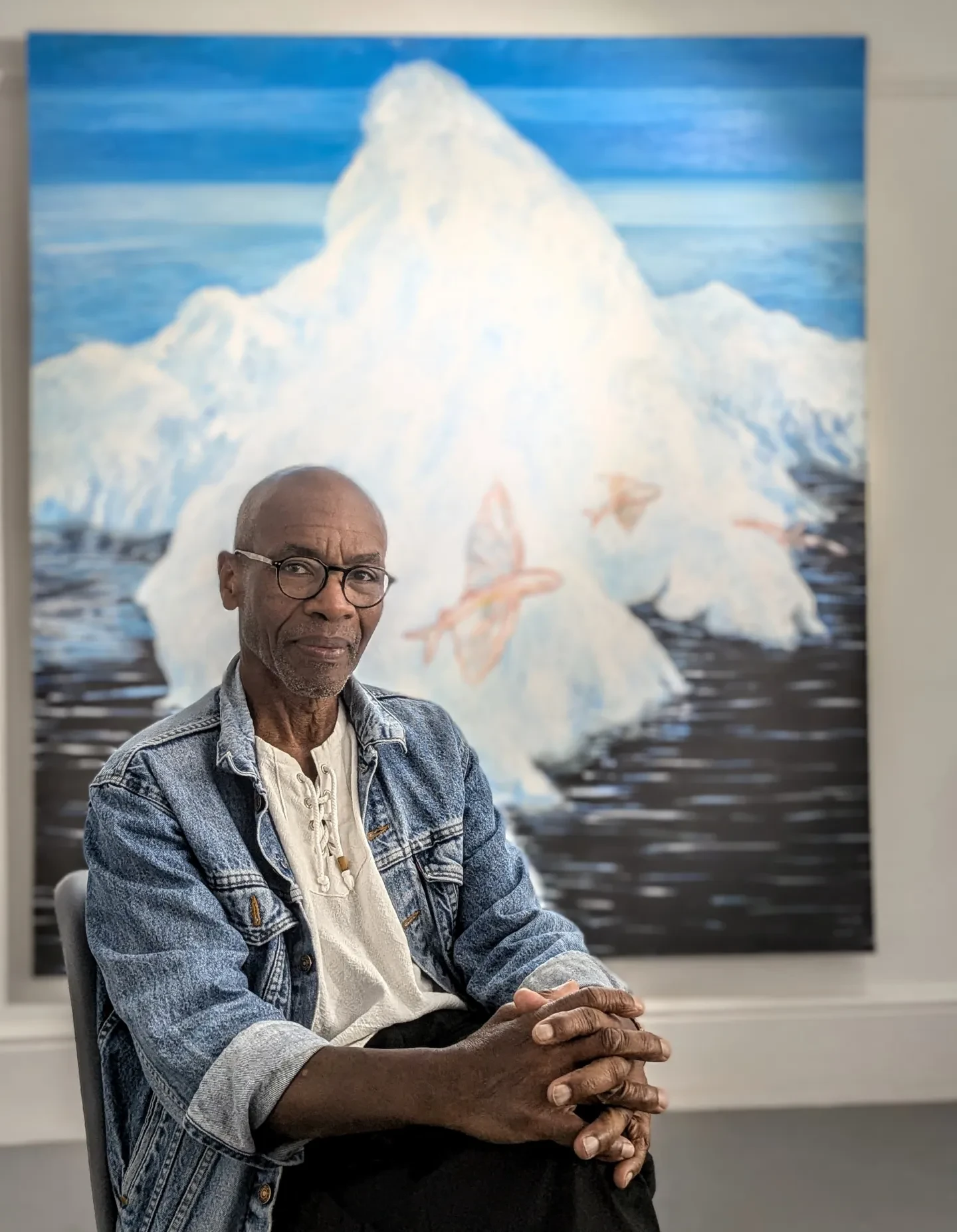 Artist Tam Joseph sits next to an artwork of an iceberg in a stormy sea with red flying fish leaping across
