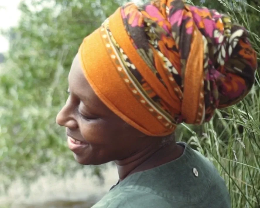 A side-on photograph of Remiiya Badru. She is smiling, with her head to one side, and wears an orange, brown and pink headscarf